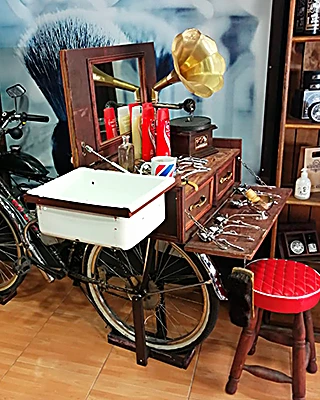 Lavabo blanco de barbería montado sobre una bicicleta antigua, con un gramófono dorado, un mueble de madera con herramientas y un banco rojo capitoné