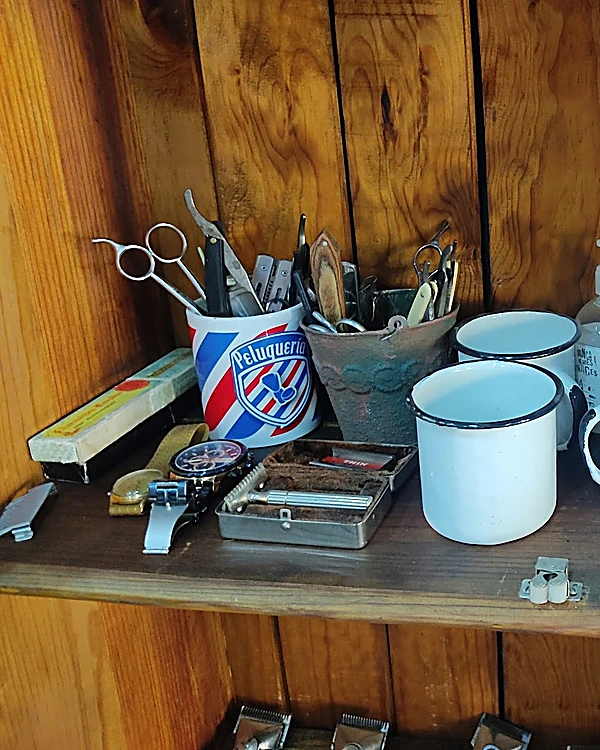 Repisa de madera con un tarro de tijeras y navajas que dice Peluquería, tazas de peltre blancas y una navaja de seguridad