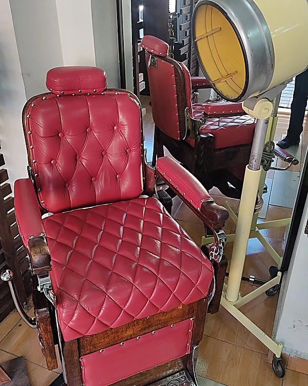 Sillón de barbero antiguo de cuero rojo capitoné con reposapiés metálico grabado, junto a una secadora de casco amarilla